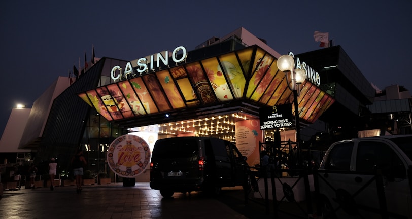 A building with a brightly illuminated entrance is seen at night. The sign above reads 'Casino' in bright letters. The facade features colorful graphics depicting various indulgent themes. Several vehicles are parked near the entrance, and people are visible in the vicinity. Tall lamp posts with spherical lights are present, casting a warm glow.