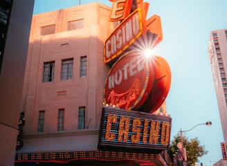 A vibrant city scene featuring a tall, illuminated casino sign with retro-style neon lights. The sign dominates the building facade with the words 'CASINO' and 'HOTEL'. Sunlight peeks through, creating a dramatic lens flare effect. The street is lively, with pedestrians casually walking and a hint of urban hustle.