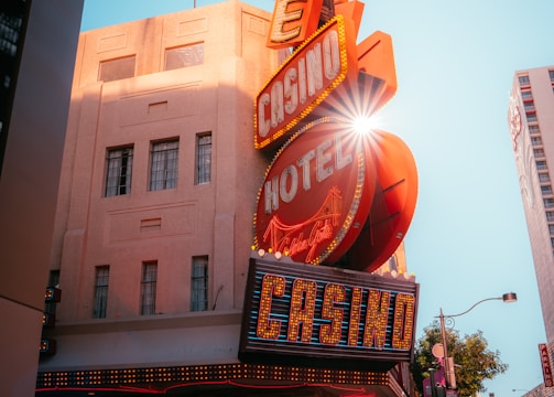A vibrant city scene featuring a tall, illuminated casino sign with retro-style neon lights. The sign dominates the building facade with the words 'CASINO' and 'HOTEL'. Sunlight peeks through, creating a dramatic lens flare effect. The street is lively, with pedestrians casually walking and a hint of urban hustle.